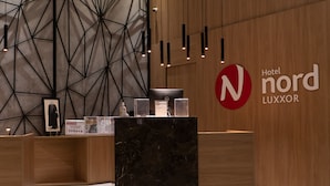 Un mostrador de recepción de hotel con diseño moderno, que presenta un patrón geométrico en la pared, una zona de recepción de madera y un letrero con el nombre Hotel Nord LUXXOR.