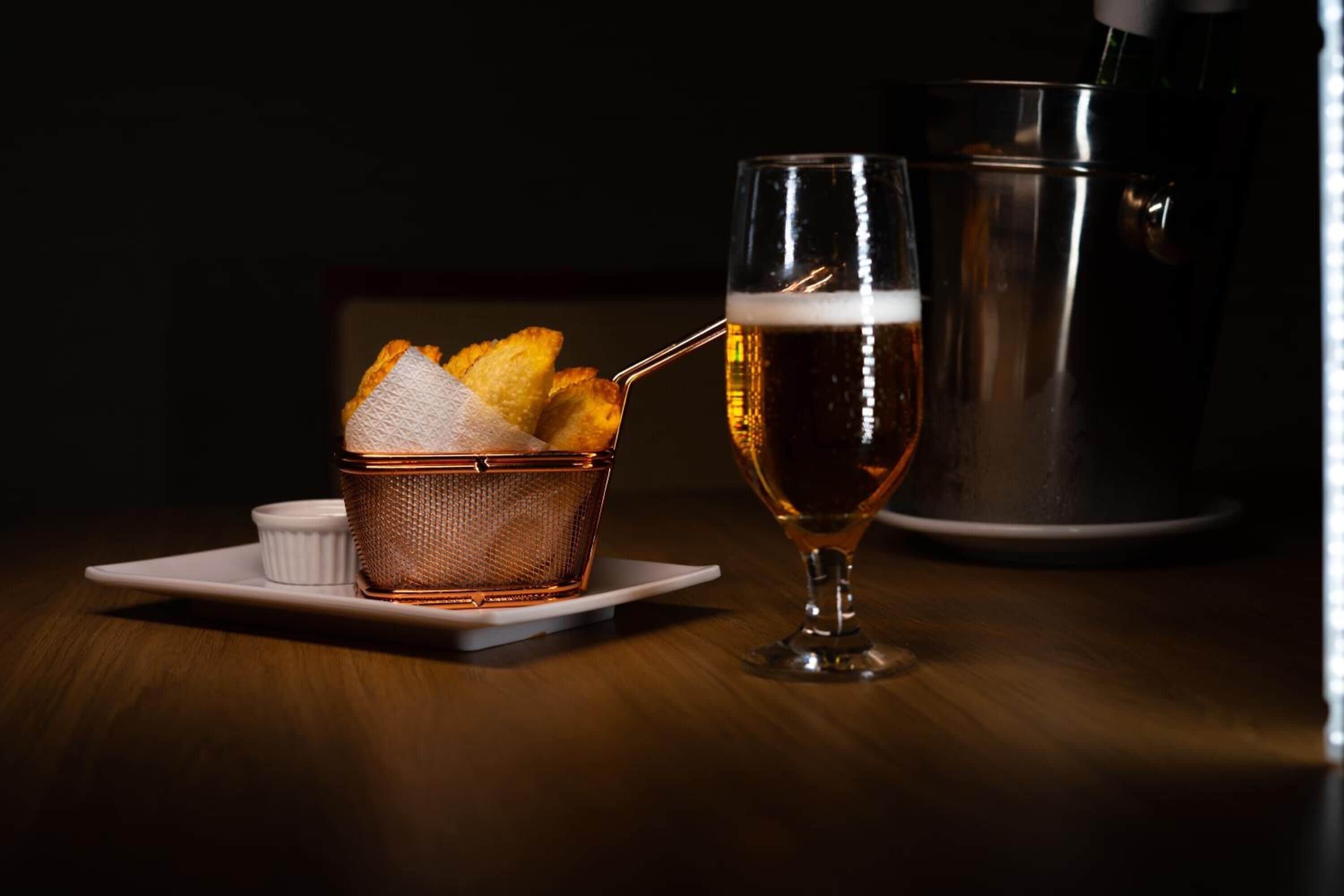 Una cerveza con pajita, una cesta de palitos de pan y un recipiente blanco pequeño sobre una mesa de madera.
