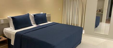 Una cama bien hecha con una colcha azul oscuro y almohadas, una mesita de noche con una lámpara y un espejo en la pared.