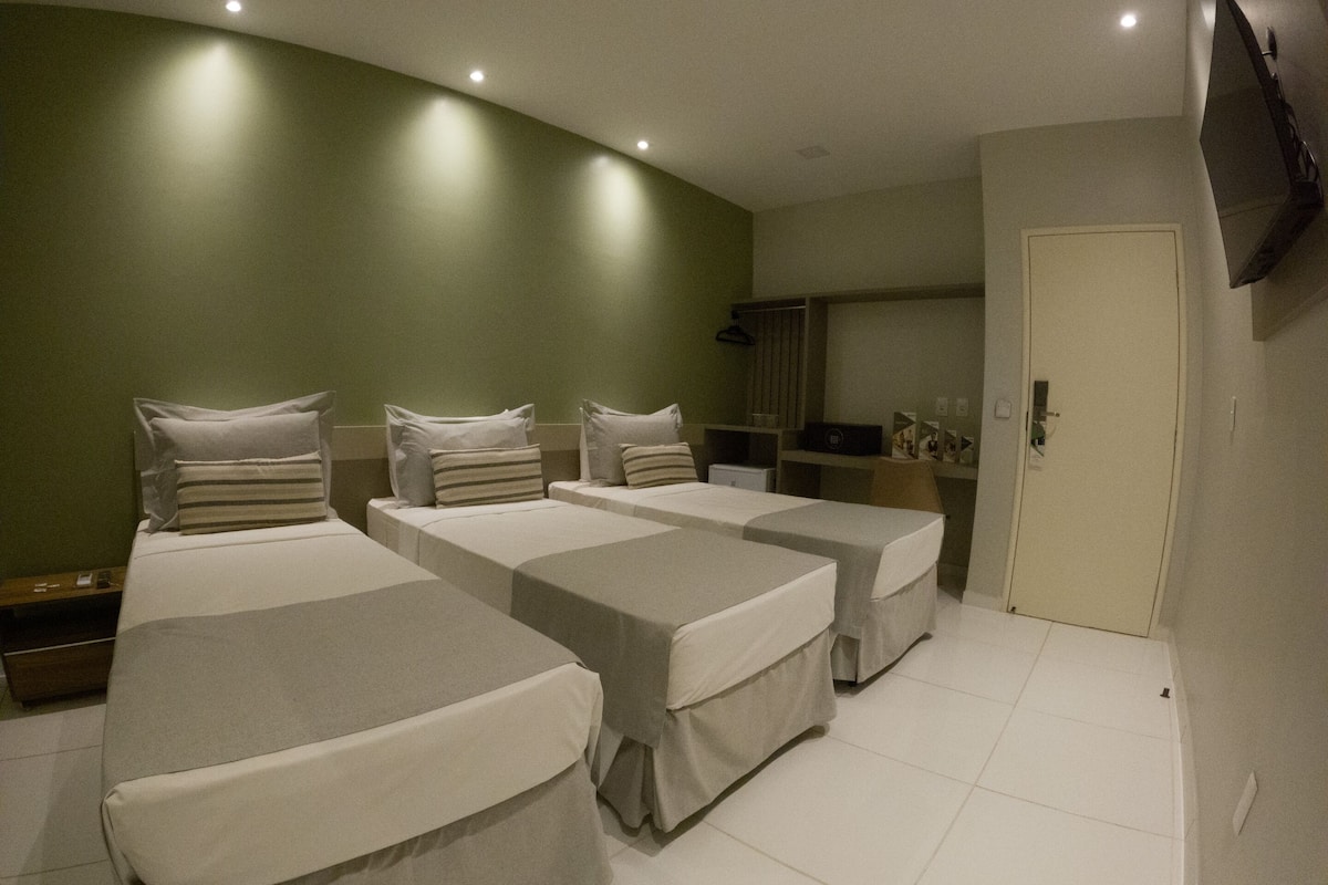 A hotel room with three single beds, a desk, a chair, a TV mounted on the wall, and a door.