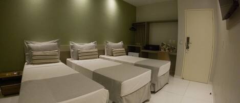 A hotel room with three single beds, a desk, a chair, a TV mounted on the wall, and a door.