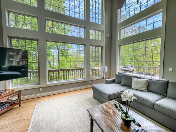 A spacious living room with large windows, a grey sofa, a coffee table with a vase, and a TV on a wooden stand. - Amazing Central Location! Lakefront! Hot tub/dock! (Oakland)