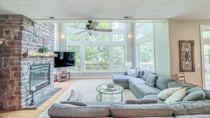 A spacious living room with a stone fireplace, a large grey sectional sofa, a glass coffee table, and a flat-screen TV mounted on the wall. - Amazing Central Location! Lakefront! Hot tub/dock! (Oakland)