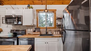 A modern kitchen with white cabinets, stainless steel appliances, a wooden ceiling, and a window with a view of trees. - SUPER CUTE & CLEAN MODERN HOME 40 min. to YNP (Island Park)