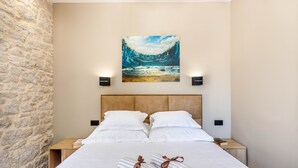 Une chambre d’hôtel avec un mur en pierre, un grand lit avec du linge de lit blanc, deux tables de chevet, un tableau représentant une vague et deux lampes fixées au mur.
