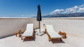 Deux chaises longues avec des structures en bois et des coussins blancs, un parasol noir et de petites tables avec des fruits et des boissons.