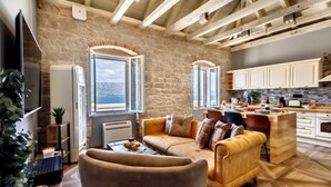 Un salon chaleureux avec un canapé et une table basse, une cuisine dotée de meubles en bois et d’un mur en pierre, et de grandes fenêtres offrant une vue sur la mer.