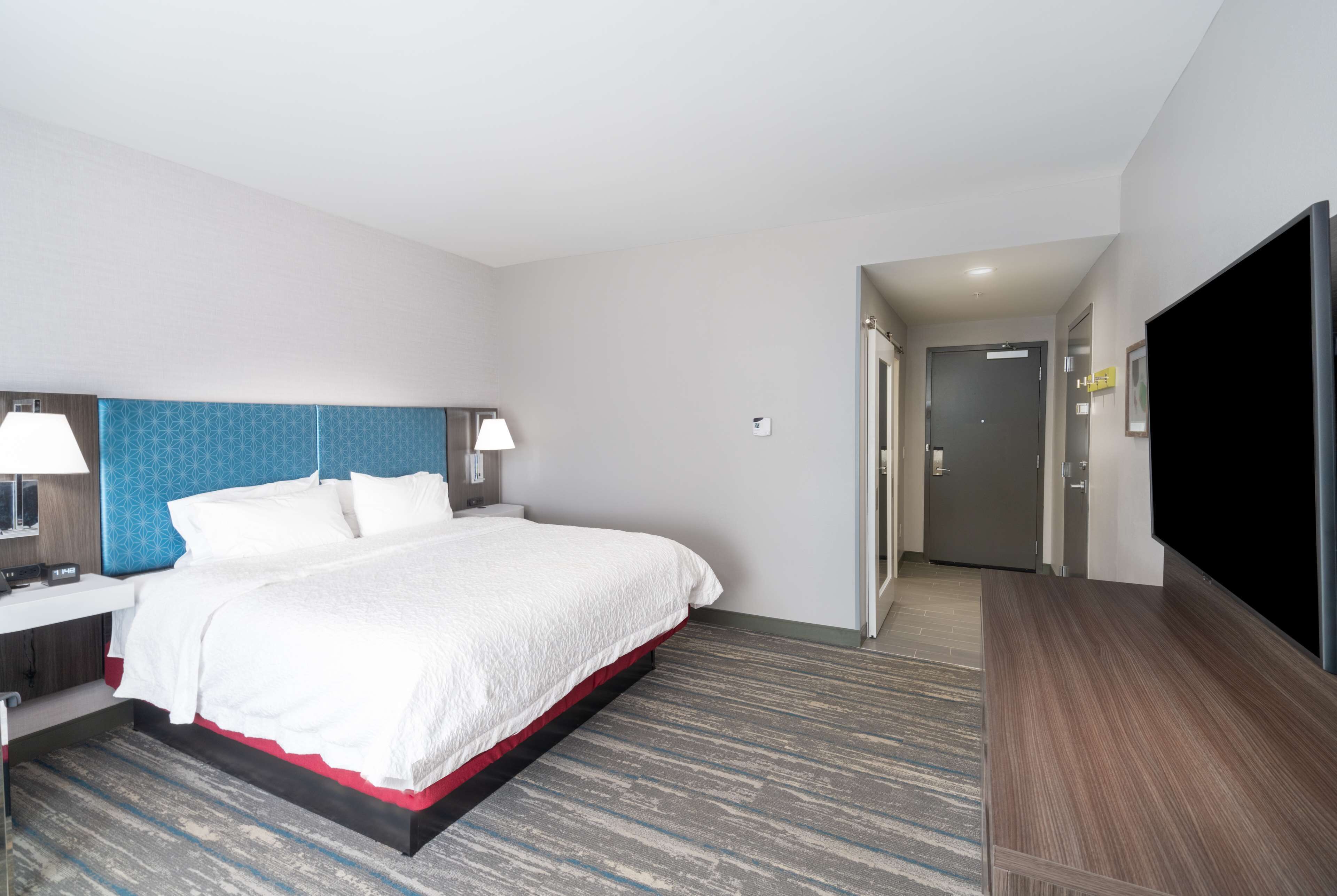 A hotel room with a large bed, bedside lamps, a flat-screen TV, and a door leading to another room.