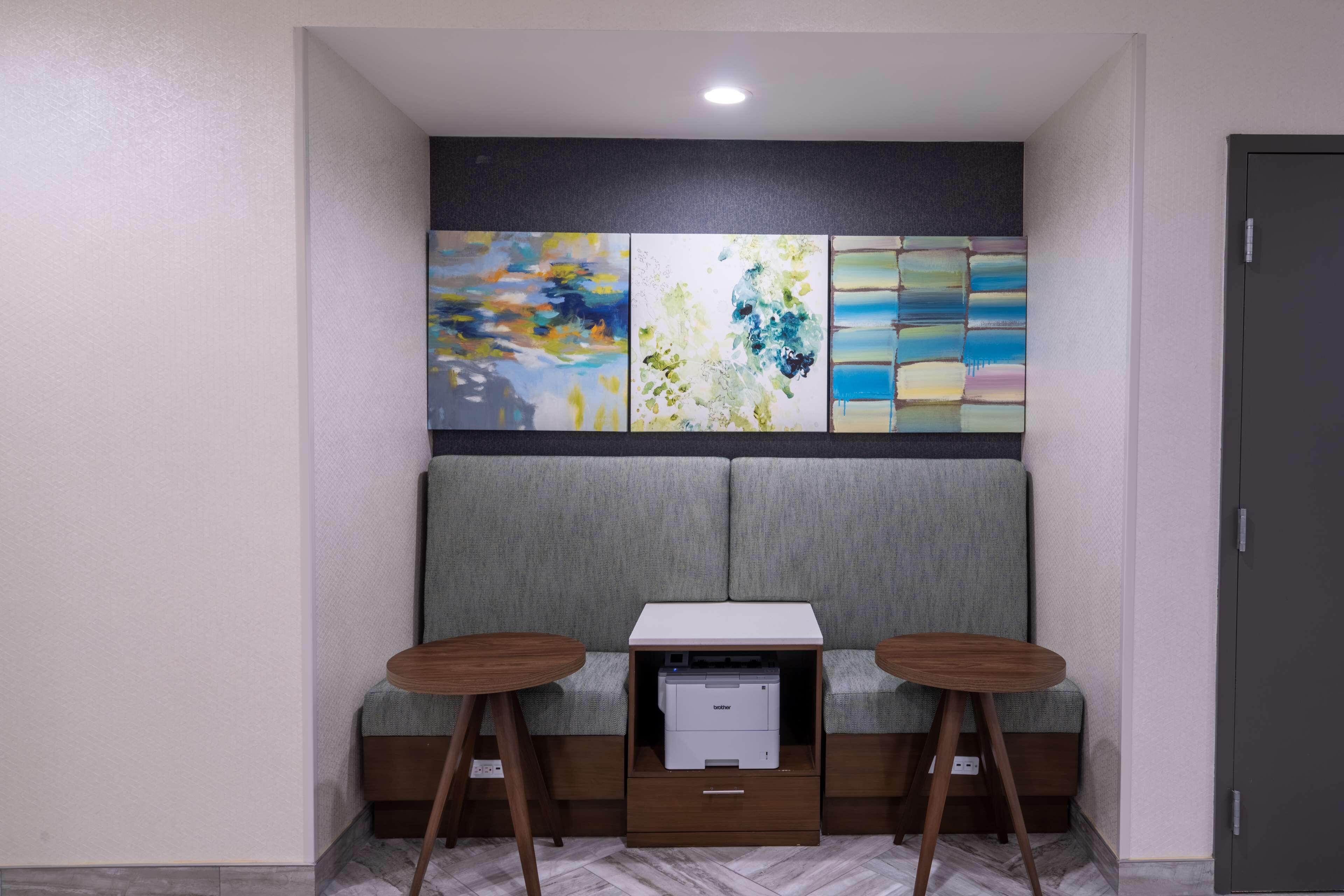 A small, modern room with a grey sofa, two wooden stools, a printer, and abstract art on the wall.