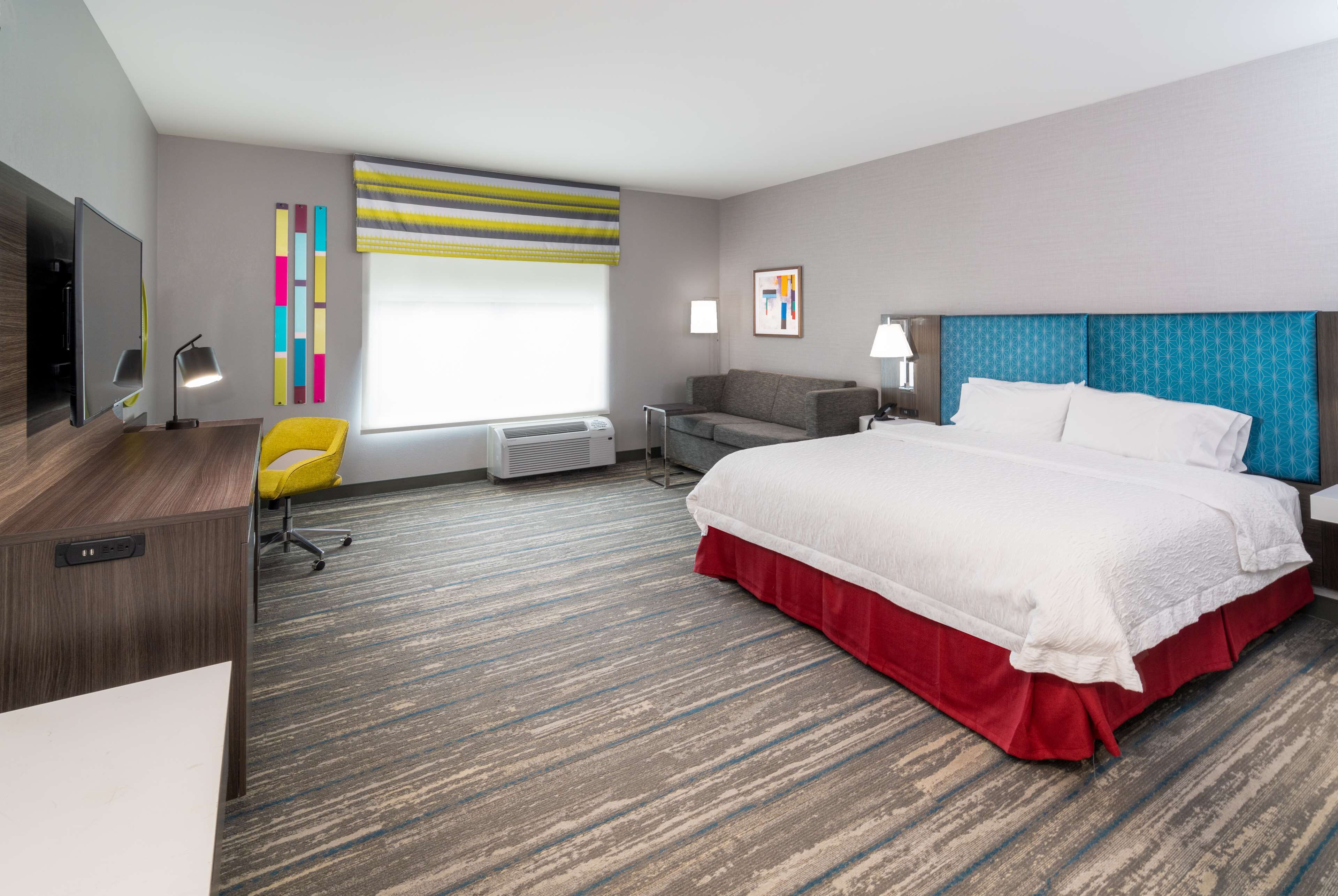 Une chambre d’hôtel avec un grand lit, un bureau, une chaise, une télévision, une fenêtre avec des rideaux et un mur orné d’une œuvre d’art colorée.
