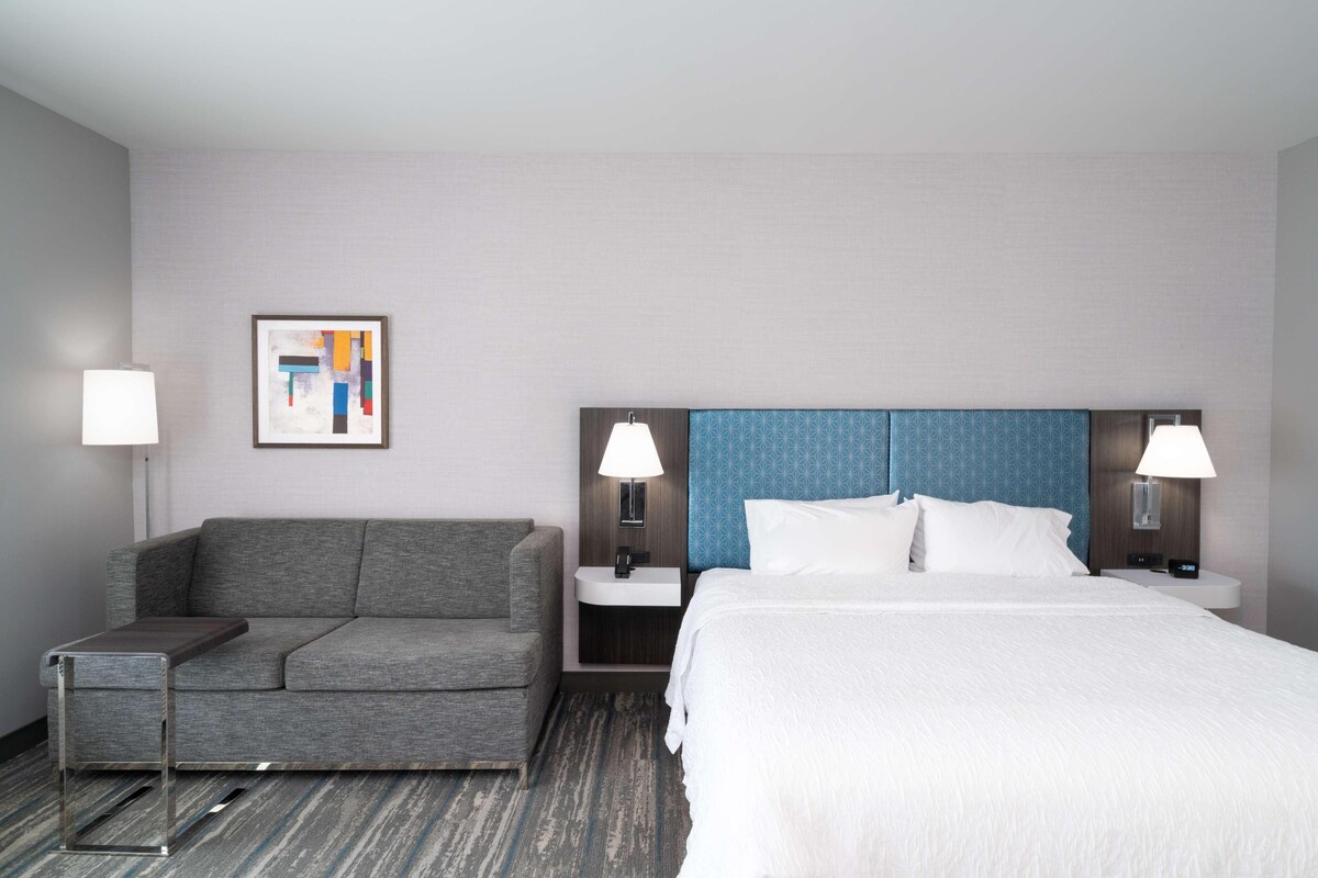A hotel room with a grey sofa, a bed with white bedding, a small bedside table, a nightstand with a lamp, and a framed picture on the wall.