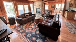 A cozy cabin interior with a living room, fireplace, wooden floors, and large windows. - Ski Access | Hot Tub | 2 King Beds | Fireplace (Whitefish)