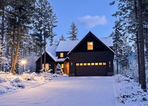 A cabin with a dark exterior, a sloped roof, and a garage with illuminated windows. - Ski Access | Hot Tub | 2 King Beds | Fireplace (Whitefish)
