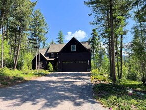 Une cabane aux couleurs sombres, dotée d’un garage, entourée de grands arbres et surplombant un ciel dégagé.
