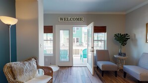 A cozy living room with a wicker sofa, patterned chairs, a lamp, and a 'WELCOME' sign above the door leading to a balcony. - Shared Pool and Hot Tub, Golf Cart Access (Port Aransas)