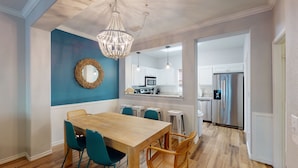 A dining area with a wooden table and teal chairs, a chandelier, and a kitchen in the background. - Shared Pool and Hot Tub, Golf Cart Access (Port Aransas)