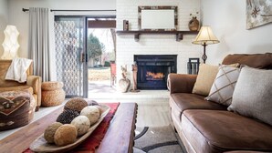 A cozy living room with a fireplace, a sofa, a coffee table with bread, and a view of the outdoors.
