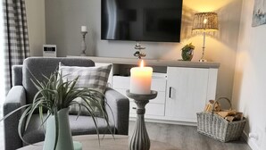 Un salon moderne avec un canapé gris, un meuble blanc, un téléviseur à écran plat, une bougie allumée posée sur un piédestal, une plante en pot et un panier en osier rempli de bûches.