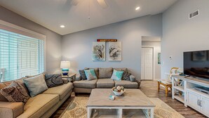A living room with a sofa, coffee table, and TV stand. There are two wall-mounted paintings and a ceiling fan. - 2nd Floor, Shared Pool, Pet Friendly, Golf Cart Access (Port Aransas)