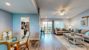 A spacious living area with a dining table, chairs, a sofa, and a coffee table. There is a kitchen area with cabinets and a microwave. A ceiling fan is visible, and the room has large windows. - 2nd Floor, Shared Pool, Pet Friendly, Golf Cart Access (Port Aransas)