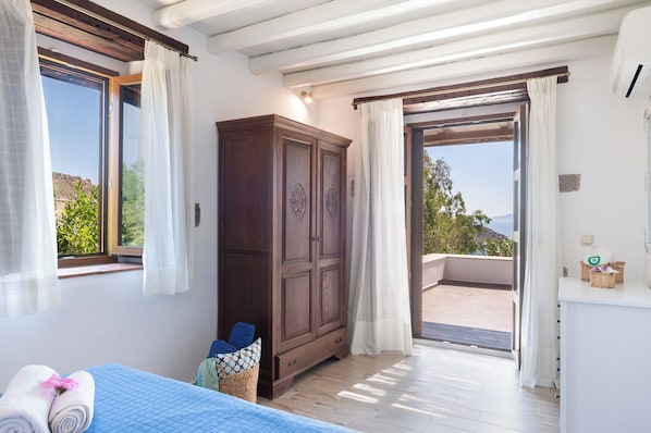 Um quarto com um guarda-roupa espaçoso, uma cama com colcha azul, uma janela com cortinas e uma varanda com vista para o mar.