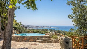 Una piscina de piedra con escalones, rodeada de árboles y una valla de madera, con vista a una ciudad costera.
