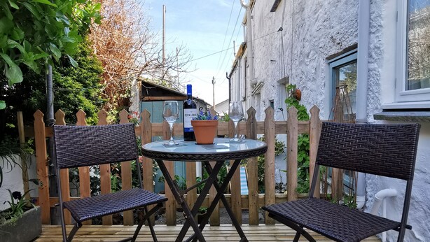 A wooden deck with a round table and four chairs, surrounded by a wooden fence and greenery.