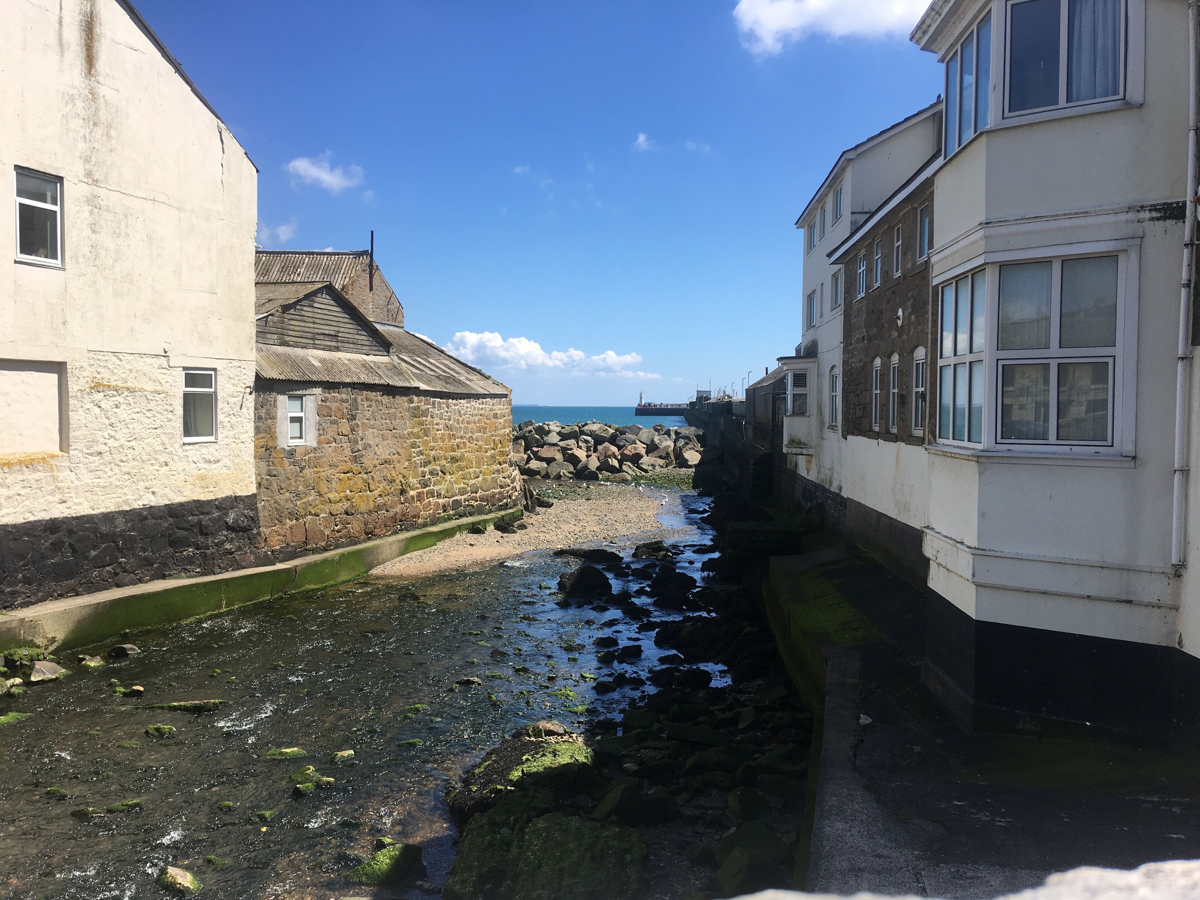 A narrow waterway between buildings with a rocky shore and a pier in the distance.