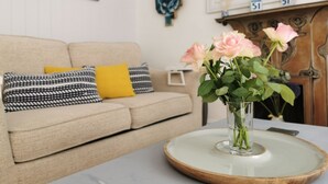 A living room with a beige sofa, patterned pillows, a vase with pink roses, and a fireplace with decorative items.