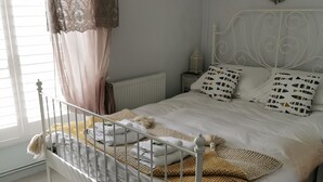 A bedroom with a white metal bed frame, a white duvet, patterned pillows, a knitted throw, and a window with curtains.