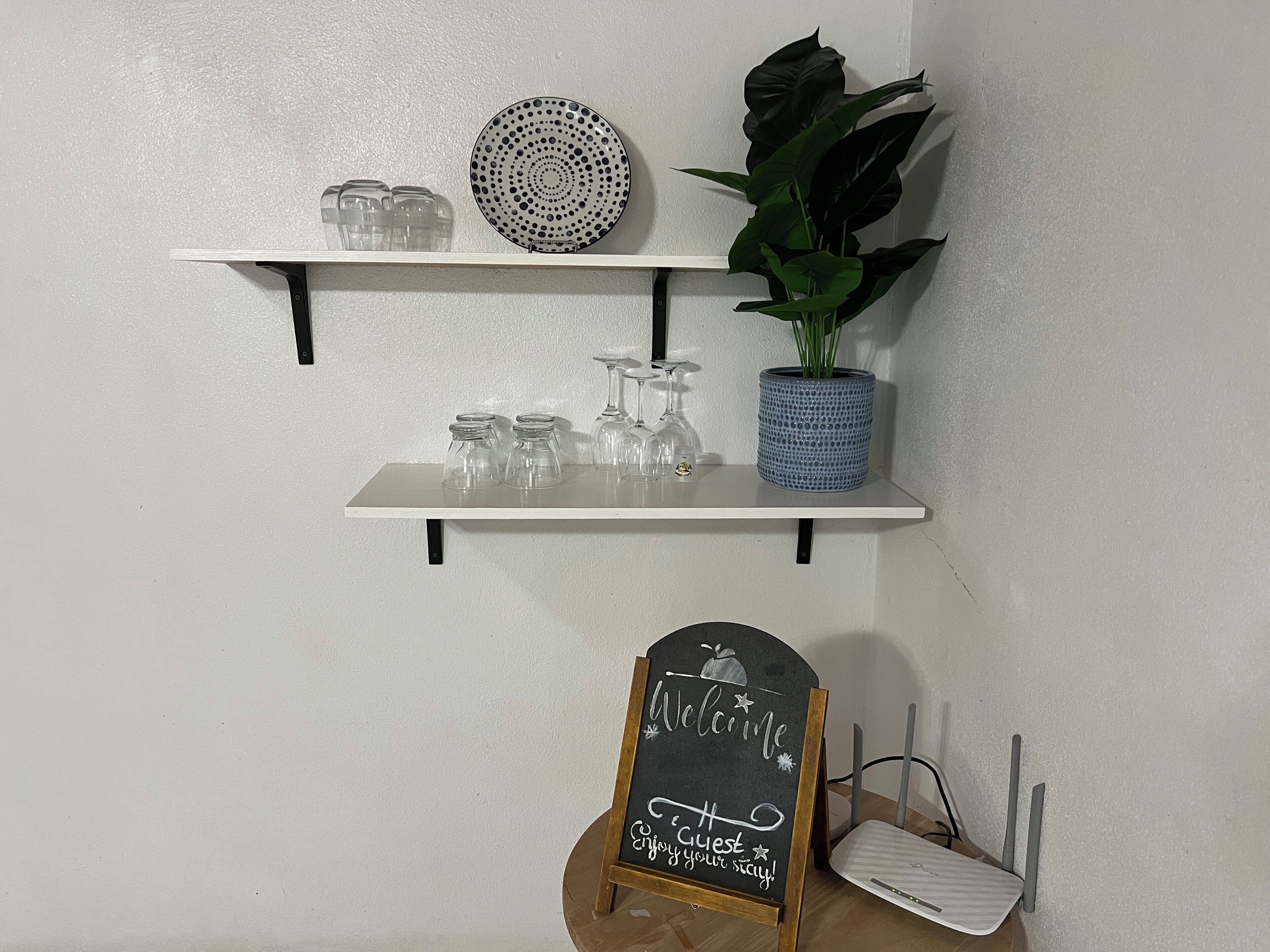 A white wall with two shelves; one holds glassware and a decorative plate, the other a potted plant and a blue vase. A chalkboard sign reads "Welcome, Enjoy your stay!"