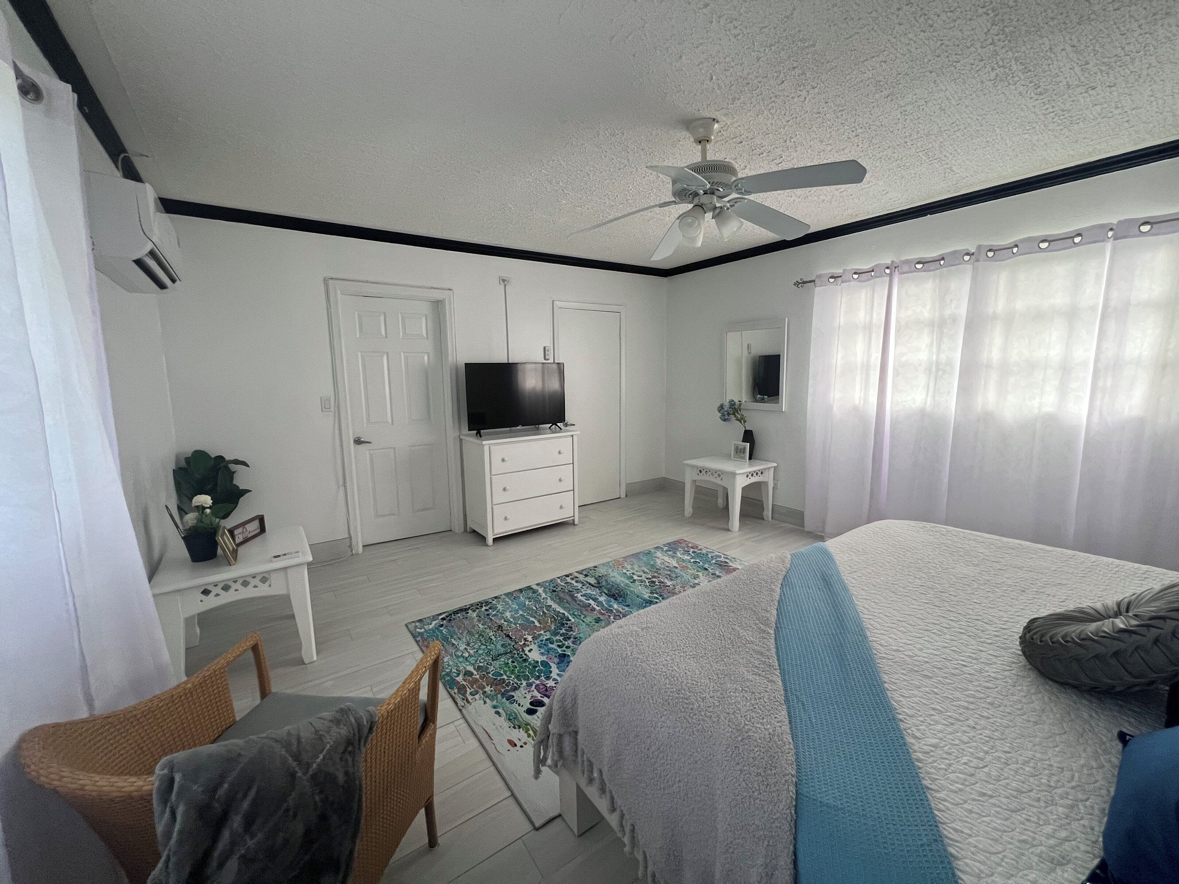 Une chambre à coucher comprenant un lit, un téléviseur, une commode, une chaise, une petite table et un ventilateur de plafond.