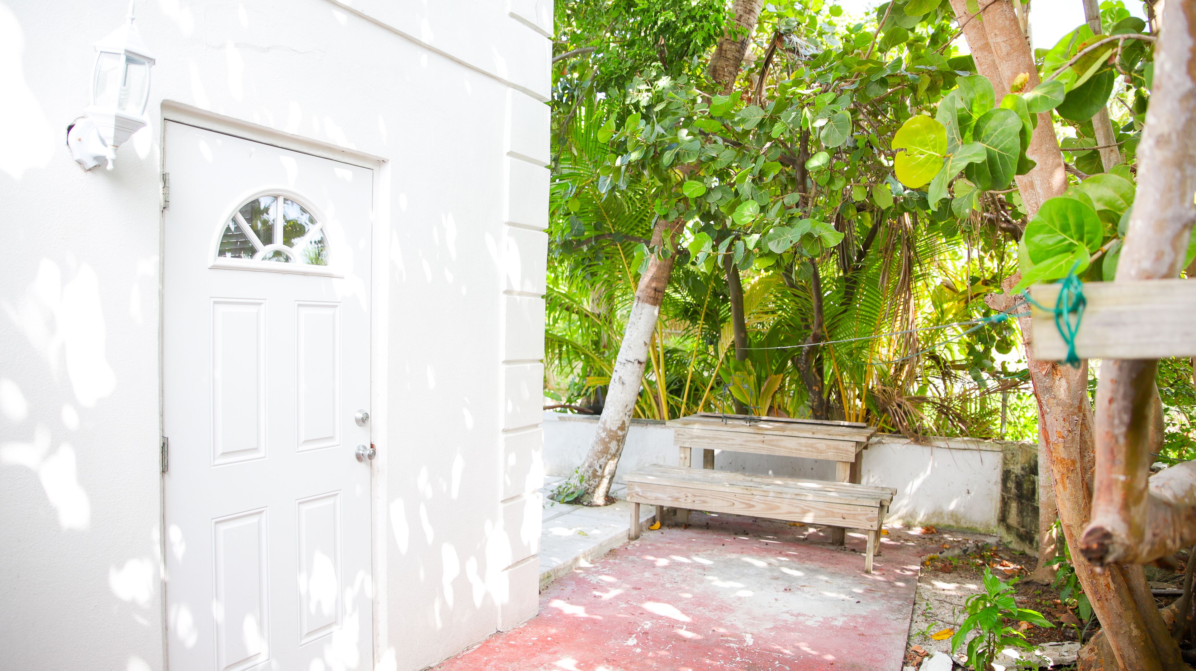 A white door with a small window, a wooden bench, and a concrete path leading to a lush green garden.