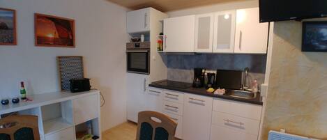 A compact kitchen with white cabinets, a microwave, a toaster, and a sink. There is a chair and a small table with a bottle and a bowl on it.