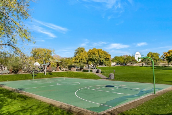 Un terrain de basket en plein air, sous un ciel dégagé, entouré d’arbres et avec un bâtiment en arrière-plan.