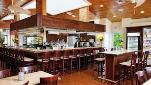 A spacious bar area with multiple seating options, a kitchen in the background, and a view of greenery outside.