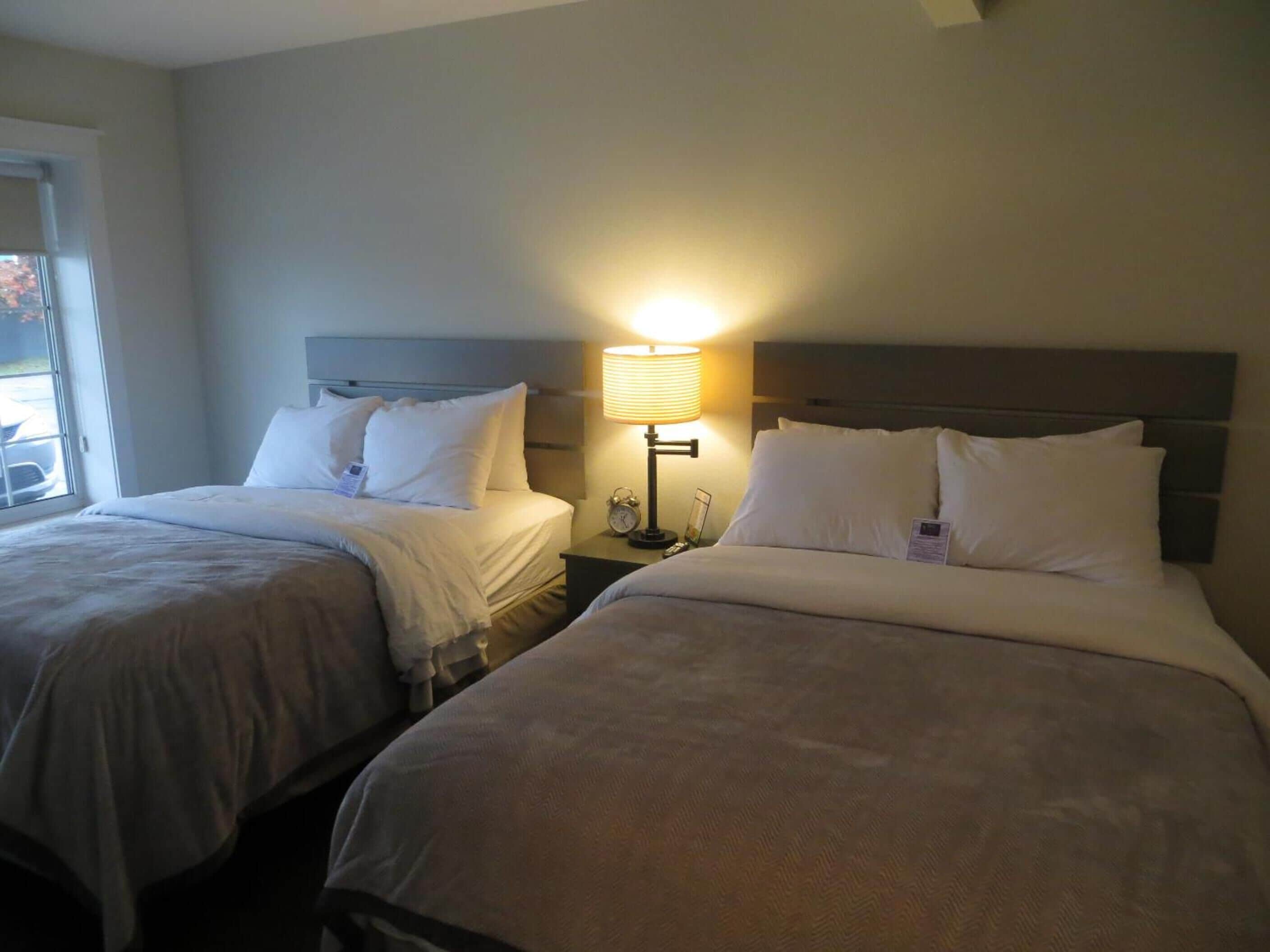 A hotel room with two beds, each with a headboard, a bedside table with a lamp, and a window with blinds.