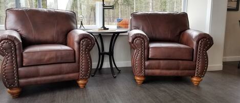 Two brown leather armchairs with wooden legs, positioned on a wooden floor with a view of trees through a window.