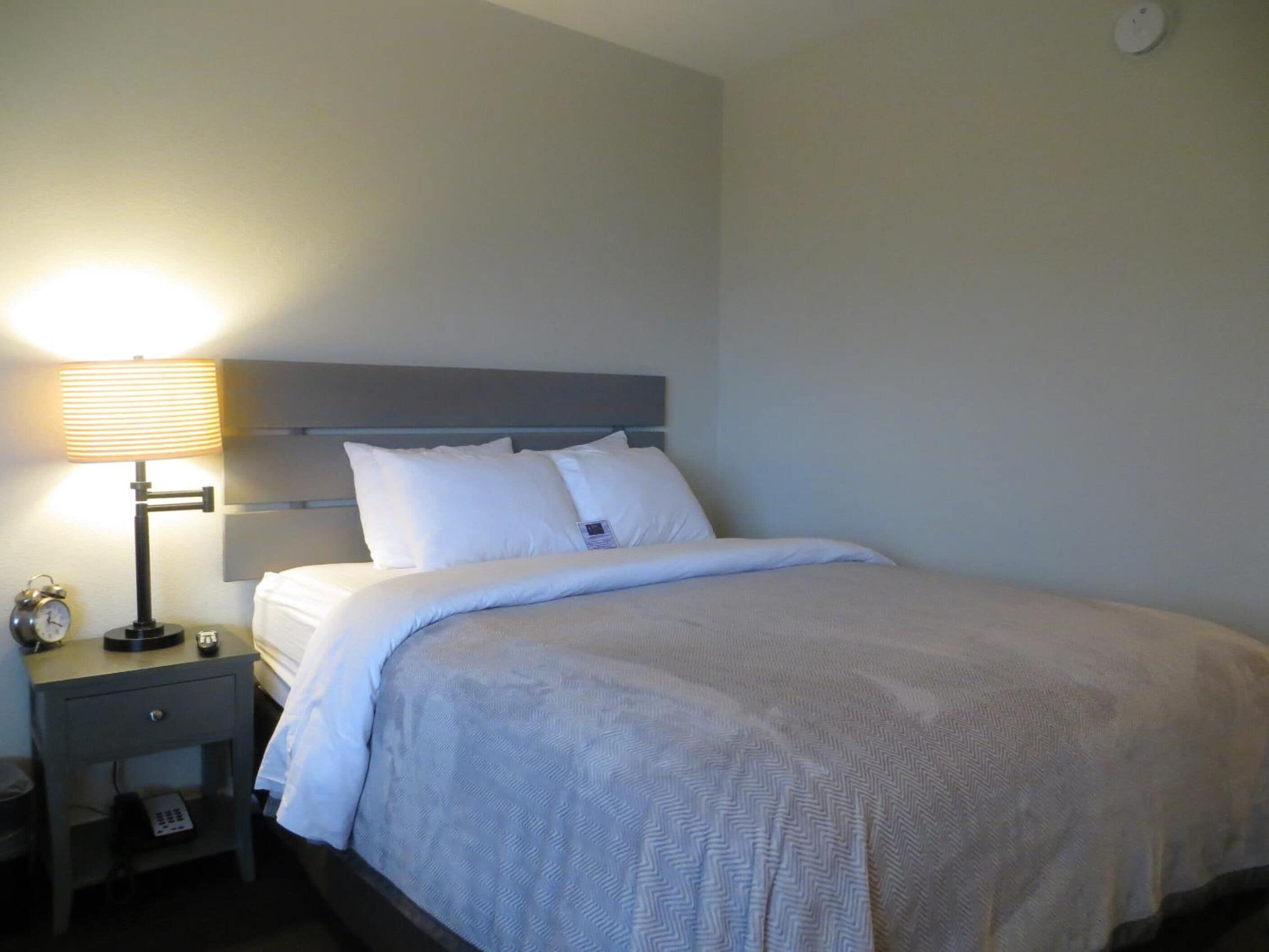 A neatly made bed with a grey headboard, a bedside table with a lamp, and a small clock.