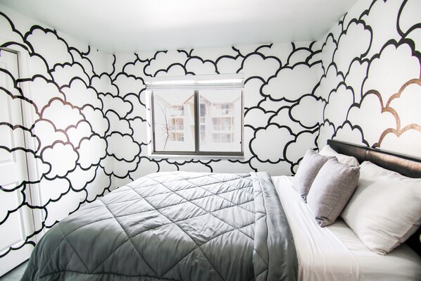 Un dormitorio con cama, ventana con vista a edificios y papel tapiz estampado con motivos de nubes.
