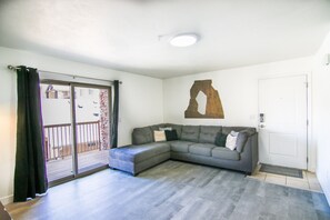 Una sala de estar con un sofá modular gris, una pared de acento de madera, un ventanal corredero y una puerta blanca.