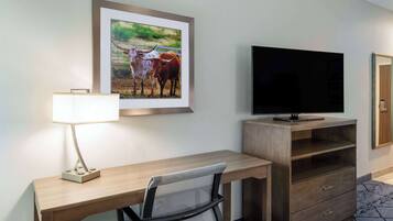 A hotel room with a wooden desk, a TV, a framed picture of cattle, and a lamp.
