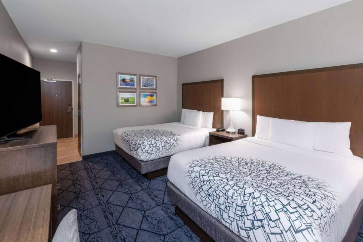 A hotel room with two beds, a TV, a desk, and a wall with framed pictures.
