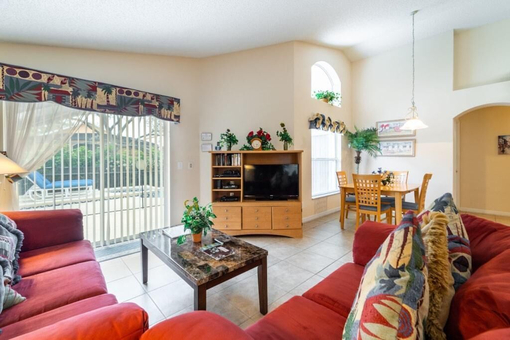 A living room with red sofas, a TV stand, a dining area with a table and chairs, and a balcony with a view.