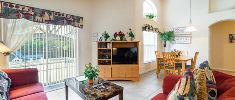 A living room with red sofas, a TV stand, a dining area with a table and chairs, and a balcony with a view.