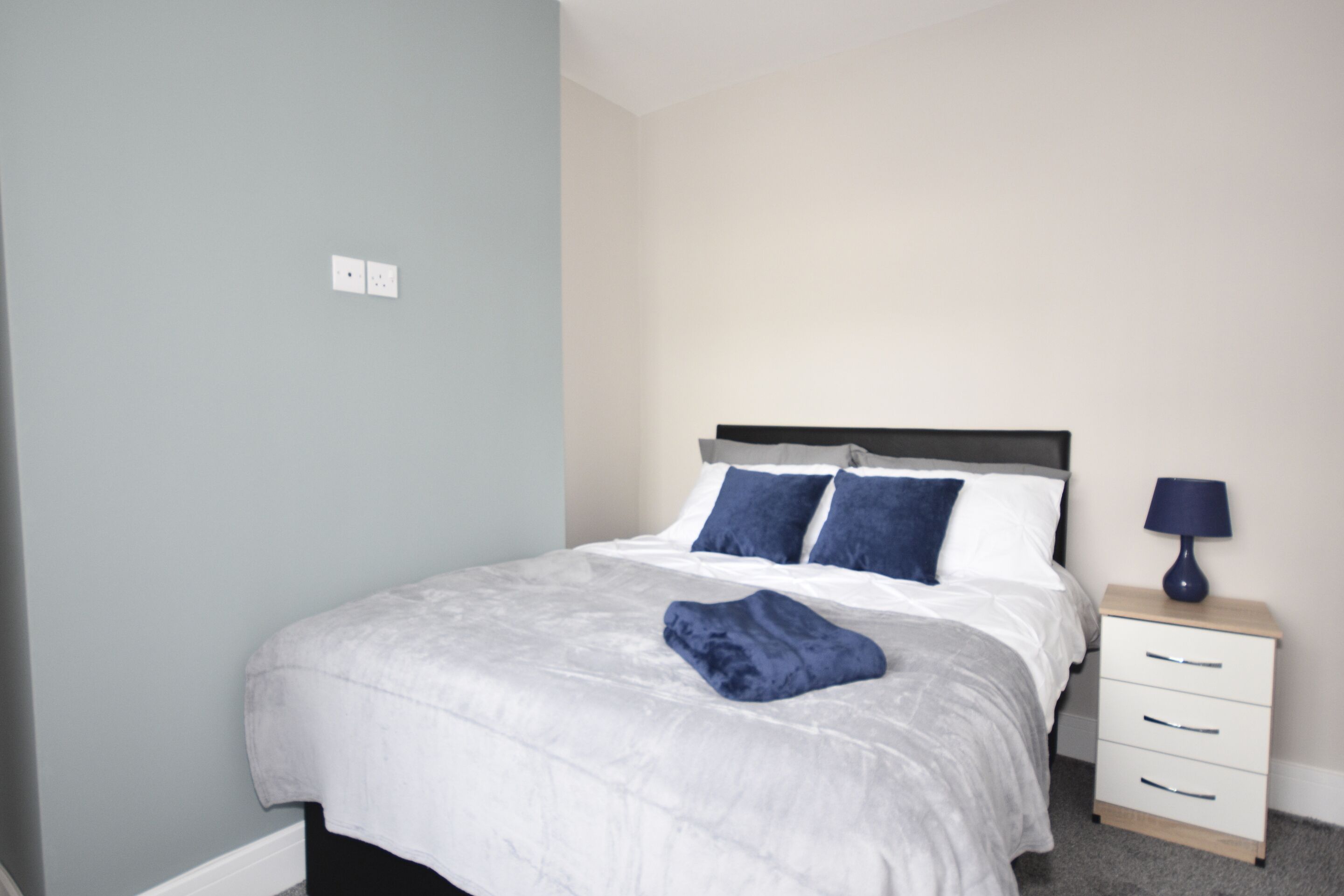 Un lit bien fait, avec une couette blanche et des oreillers bleus, une table de chevet avec une lampe, et un mur bleu clair.
