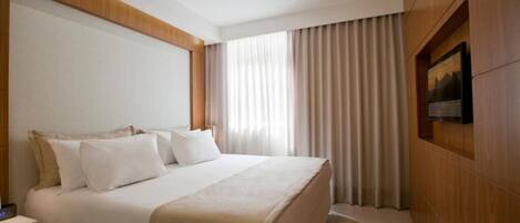 Habitación de hotel con una cama grande, una mesita de noche, un televisor de pantalla plana en la pared y una ventana con cortinas.