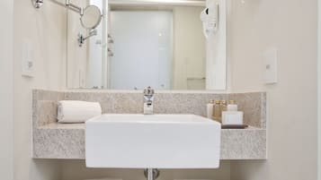 Uma casa de banho moderna com lavatório branco, bancada de granito e espelho acima. Existem dois porta-toalhas abaixo do lavatório, e um secador de cabelo fixado na parede à direita.