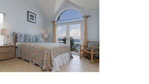 Une chambre à coucher comprenant un lit, une chaise, une table de chevet et une fenêtre donnant sur l’océan.