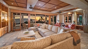 A spacious living area with a large sofa set, a coffee table, and a dining table. There is a view of palm trees and a sunset through the glass doors. - Oceanview, Private Pool and Hot Tub! (Launiupoko)